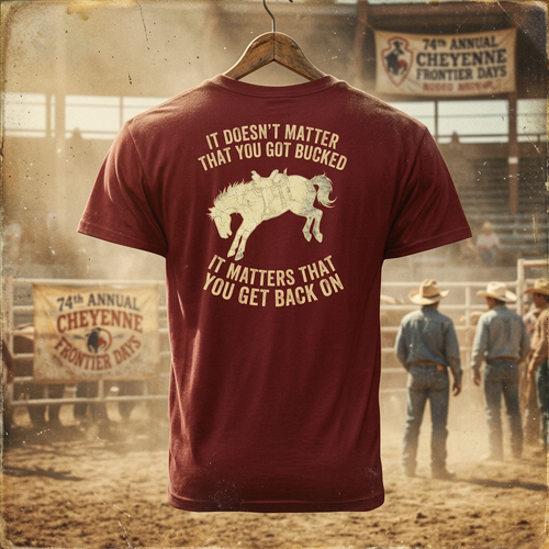 A maroon short-sleeve T-shirt hanging on a wooden hanger against a dusty rodeo arena background. The shirt features a large cream graphic of a bucking horse with the text 'IT DOESN'T MATTER THAT YOU GOT BUCKED IT MATTERS THAT YOU GET BACK ON'.