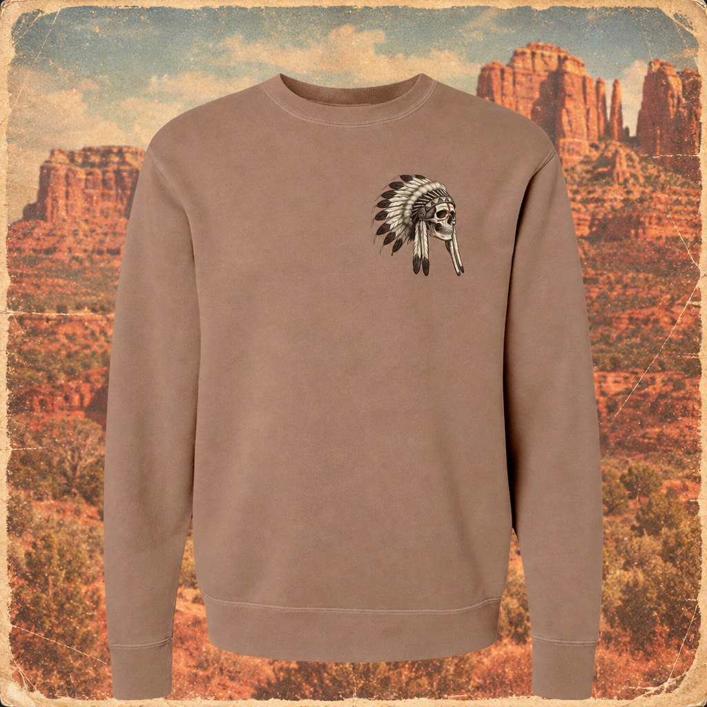 A taupe crewneck sweatshirt featuring a black and white graphic of a skull wearing a feathered headdress on the left chest, presented against a vintage-style red rock desert background.