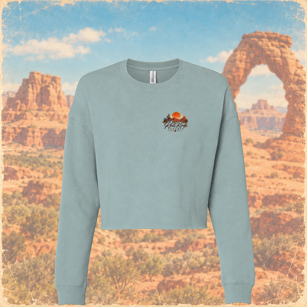 A light blue cropped crewneck sweatshirt with a small graphic on the left chest, displayed against a scenic desert background featuring Delicate Arch.